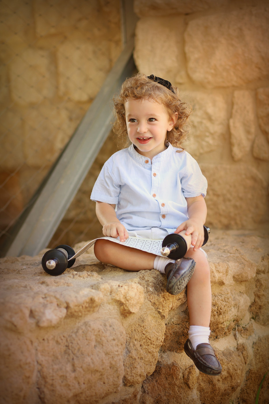 ילד מתולתל עם כיפה יושב על קיר אבן ומחזיק ספר תורה קטן
