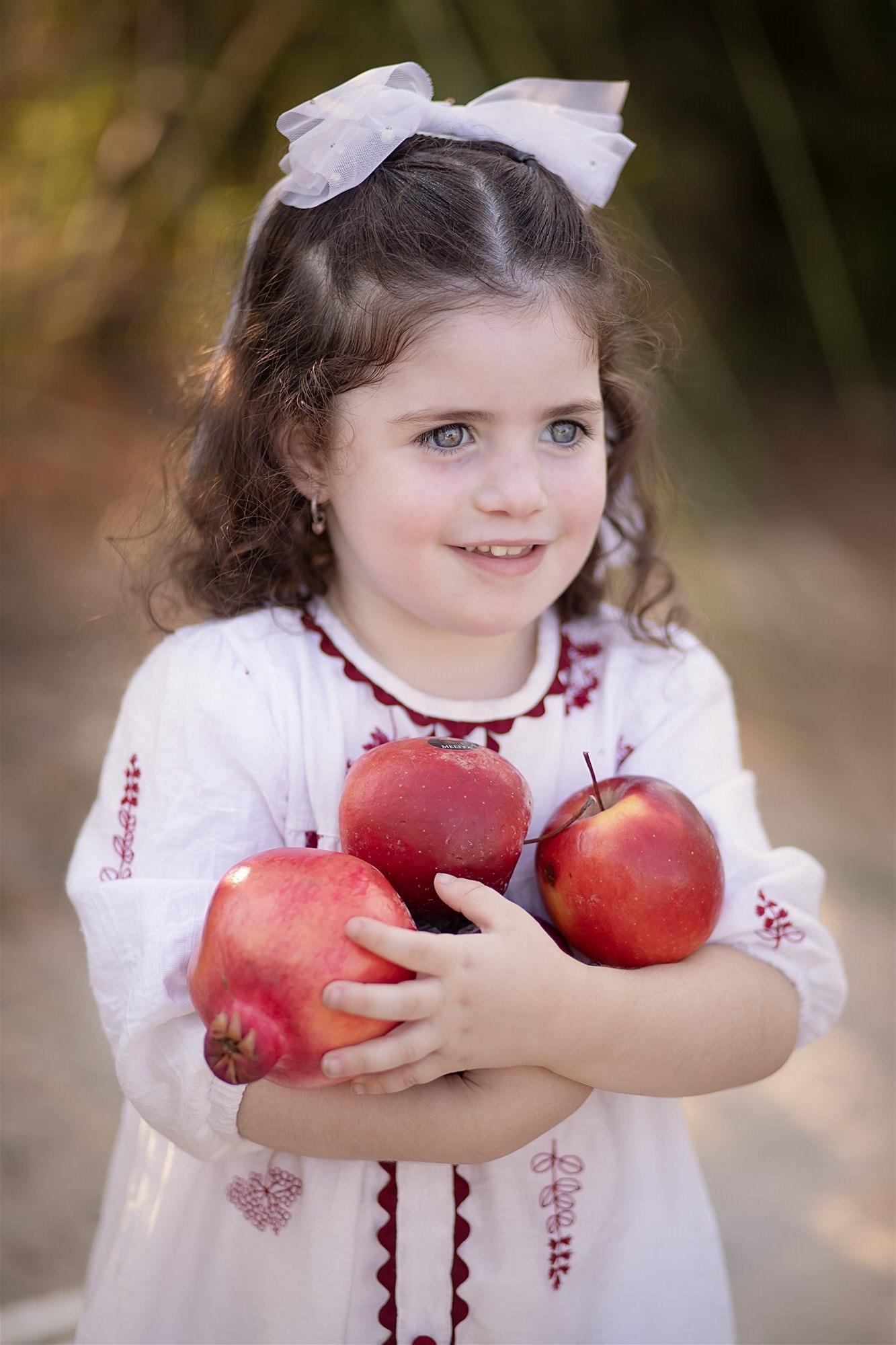 ילדה מחייכת בשמלה לבנה מחבקת תפוחים ורימון